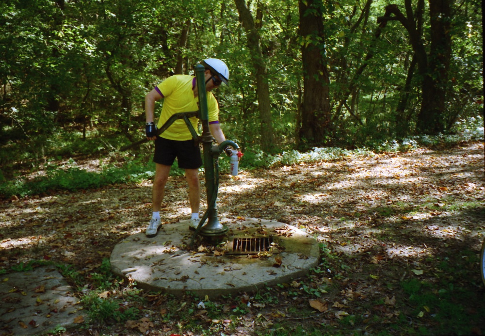 Dennis filling water bottle at well with handpump.